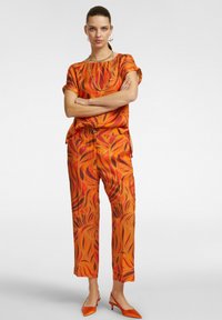 Top e pantaloni arancioni con motivo a foglia in tessuto fluido. Gli accessori includono una collana e scarpe con tacco arancioni.