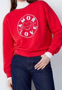 Sudadera roja con texto blanco que dice "AMOR" y "LOVE" junto con un diseño de corazón, con un cuello redondo clásico y puños acanalados.