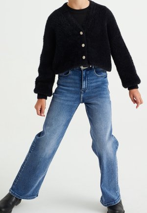 Person trägt einen schwarzen, flauschigen, geknöpften Cardigan, ein schwarzes Oberteil, hoch taillierte ausgewaschene blaue Jeans und schwarze Stiefeletten, steht mit gespreizten Beinen.