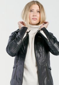 Schwarze Lederjacke mit gerippten Schulterakzenten, seitlichen Reißverschlusstaschen und einer taillierten Passform, kombiniert mit einem leichten Rollkragenpullover.
