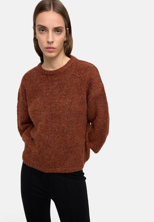 Kobieta o prostych brązowych włosach, ubrana w rdzawy, teksturowany sweter i czarne spodnie, stojąca na jasnoszarym tle.