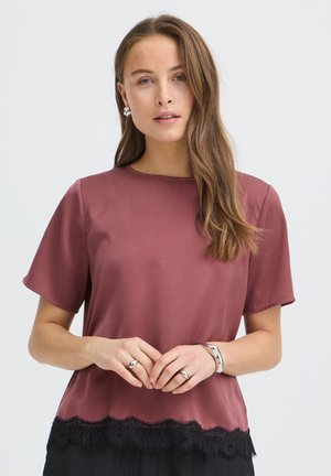 Kobieta o długich brązowych włosach, ubrana w krótki top w kolorze mauve z czarną koronkową lamówką, srebrne kolczyki koła, pierścionki i bransoletka na jednolitym tle.