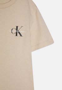 Beżowa bawełniana koszulka z krótkim rękawem, z czarnym logo "CK" i napisem "Calvin Klein Jeans" po lewej stronie. Gładka faktura.