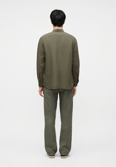 Άνδρας που στέκεται με την πλάτη σε εμάς, φοράει πουκάμισο μακρυμάνικο ελιάς και παντελόνι στο ίδιο χρώμα, μπροστά σε ένα απλό λευκό φόντο.