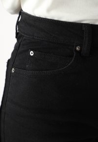 Calças de ganga preta de cintura alta com um design de cinco bolsos, apresentando ferragens metálicas e costura em contraste. O tecido tem uma textura suave.