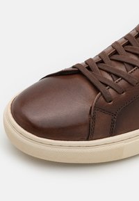 Brązowe skórzane sneakersy z zaokrąglonym noskiem, teksturowanym wierzchem i kontrastującą jasną gumową podeszwą. Wyposażone w brązowe sznurowadła i subtelne detale przeszycia.