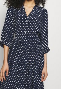 Marindräkt med vitt cirkulärt mönster, långa ärmar, knappfront och plisserad kjol. Tillverkad av lättviktig tyg med krage.