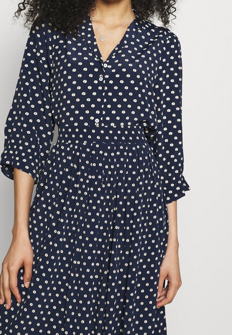 Marindräkt med vitt cirkulärt mönster, långa ärmar, knappfront och plisserad kjol. Tillverkad av lättviktig tyg med krage.