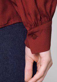 Punainen, teksturoitu paita, jossa on kerrostetut hihat ja ruskea nappi hihassa, yhdistettynä tummansinisestä denimmateriaalista näkyvään osaan alla.