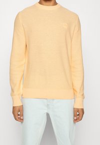 Man med en ljus persikofärgad texturerad långärmad tröja och ljusblå jeans, som står mot en enkel vit bakgrund.