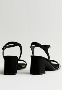 Sandali neri in suede con tacco a blocco, cinturino alla caviglia e dettagli in metallo dorato lungo il bordo. Design aperto sul retro, texture liscia.