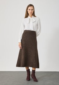 Bijela bluzica s kragnom u obliku mašne, uparena s smešnim vunenim suknjama do koljena. Model nosi burgundske gležnjače. Minimalistička pozadina.