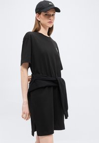 Jeune personne portant une robe T-shirt noire surdimensionnée, un sweat-shirt noir noué à la taille, une casquette noire et une petite boucle d'oreille, debout devant un fond clair.