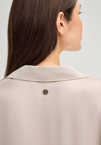Beige blouse met een klassieke kraag, zachte satijnen textuur en een decoratieve ronde metalen knop aan de achterkant met een boomontwerp.