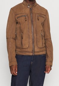 Uomo che indossa una giacca in suede marrone con cerniera, con tasche sul petto e sui lati, abbinata a jeans blu scuro, in piedi contro uno sfondo neutro.