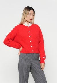 Cardigã de malha oversized vermelho com decote redondo, botões prateados e punhos canelados. Combinado com calças cinzas risca de giz.