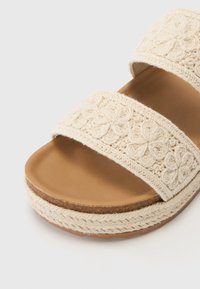 Sandale à enfiler beige en tissu tissé, ornée de deux brides à motif floral, avec une semelle plate marron et un bord tressé texturé.
