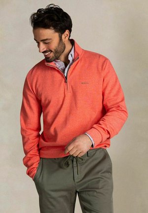 Mann trägt einen korallenroten Pullover mit Viertelreißverschluss über einem gestreiften Hemd und olivgrüne Hose mit Kordelzug, lächelt und schaut nach unten, eine Hand in der Tasche.