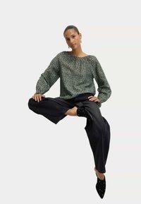 Green patterned blouse with long sleeves, paired with black wide-leg jeans and pointed-toe black heels featuring circular accents.