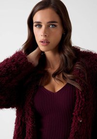 Bordeauxroter strukturierter Cardigan aus flauschigem Garn, getragen über einem eng anliegenden, gerippten Top in derselben Farbe, mit V-Ausschnitt und glattem Stoff.