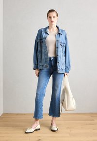Jeansjakke, ribbet beige topp, lyseblå flagrende jeans og kremfargede leiligheter med sløyfe-detalj. Holder en hvit strukturert veske. Nøytral bakgrunn.