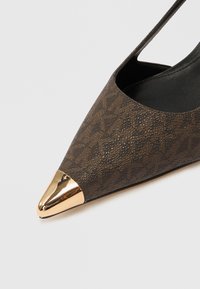 Smeđa uzorkovana cipela na visoku petu sa šiljastim vrhom i zlatnim metalnim vrhom. Ima teksturiranu površinu i otvoreni stražnji dizajn.