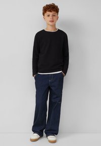 Zwarte longsleeve shirt met witte halslijn, gecombineerd met losse blauwe denim jeans en witte sneakers met tan kleurige zolen, staande tegen een neutrale achtergrond.