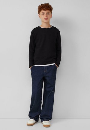 Schwarzes Langarmshirt mit weißem Halsausschnitt, kombiniert mit lockeren blauen Denim-Jeans und weißen Turnschuhen mit braunen Sohlen, vor einem neutralen Hintergrund stehend.