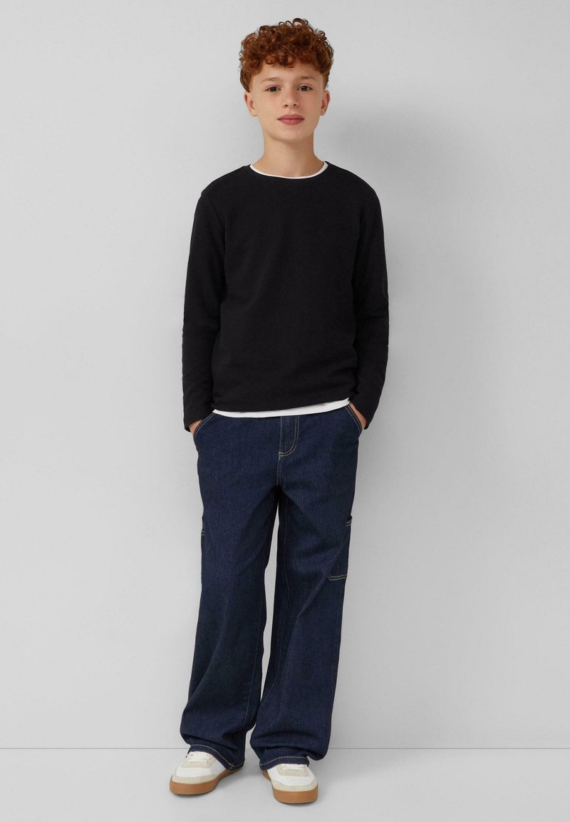 Zwarte longsleeve shirt met witte halslijn, gecombineerd met losse blauwe denim jeans en witte sneakers met tan kleurige zolen, staande tegen een neutrale achtergrond.