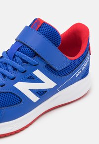 Blå sportskor med vit logotyp, meshmaterial och röda accenter. Har en kardborrestrap och strukturerad sula för grepp.