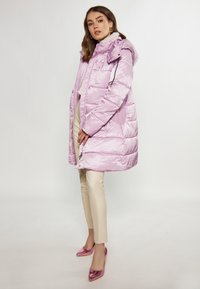 Geacă puf light purple cu glugă, finisaj lucios, design matlasat, buzunare laterale și accente negre pe șnur, asortată cu pantaloni crem și tocuri roz.