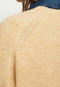 Pull en maille beige avec une finition texturée, doté de manches raglan et d'un col rond, superposé sous un col en denim foncé.