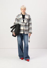 Cappotto a quadri grigio e bianco con dettagli marroni, design monopetto e ampi revers. Maglione scuro e jeans in denim svasati. Borsa a puffo nera. Scarpe rosse.