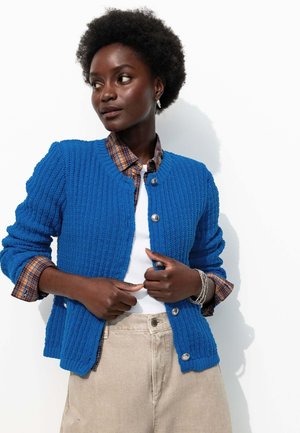 Cardigan en maille bleu avec un motif texturé, fermeture à boutons et poignets côtelés, porté sur une chemise à carreaux et un haut blanc, sur fond blanc.
