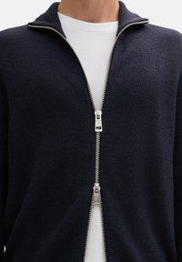 Granatowy cardigan na zamek błyskawiczny wykonany z teksturowanej dzianiny, z prążkowanym kołnierzem i podwójnym srebrnym wykończeniem zamka, noszony na białej koszuli.