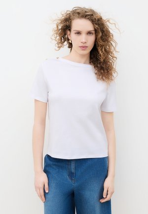 Junge Frau mit lockigen Haaren, die ein weißes kurzärmeliges Oberteil mit Knöpfen und blaue Jeans trägt, steht vor einem schlichten weißen Hintergrund.