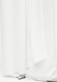 Λευκό αέρινο φόρεμα καλύπτει λευκά μυτερά παπούτσια, αναδεικνύοντας την υφή του υφάσματος και ελαφριά κίνηση στο στρίφωμα.