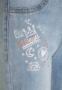 Jeans de mezclilla azul claro con bordados decorativos en blanco y oro rosa, que incluyen motivos de una corona, corazones, lunas y texto.