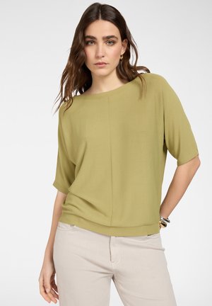 Donna con capelli castano ondulati, indossa una maglietta a maniche corte verde oliva e pantaloni beige chiaro, posando con una mano sul fianco davanti a uno sfondo semplice.