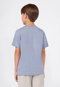 Niño con cabello castaño corto, vistiendo una camiseta de color azul claro y pantalones beige, de espaldas contra un fondo blanco.