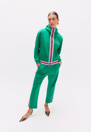 Mujer con chándal verde con franjas rojas, blancas y azules, de pie con las manos en los bolsillos y calzado con punta y estampado.