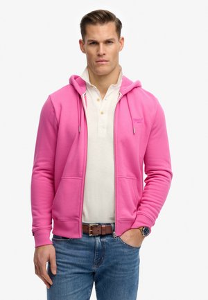 Mann trägt ein weißes Hemd mit Knöpfen, einen pinkfarbenen Kapuzenpullover mit Reißverschluss, blaue Jeans, braunen Gürtel und eine Armbanduhr und steht mit einer Hand in der Tasche.