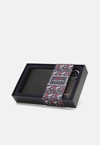Portefeuille noir dans une boîte cadeau avec un ruban de vacances à motifs. La boîte présente un intérieur bleu marine et une étiquette indiquant "Joyeuses Fêtes".