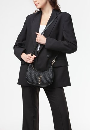 Donna che indossa un completo nero e top bianco tiene una borsa a tracolla nera testurizzata con logo V73 dorato sul davanti.