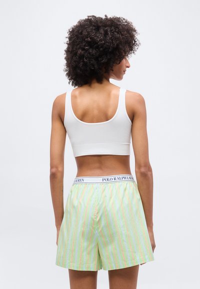 Γυναίκα με σγουρά μαλλιά, που φοράει λευκό κοντό τοπ και ριγέ σορτς Polo Ralph Lauren σε παστέλ αποχρώσεις, φωτογραφημένη από πίσω με απλό φόντο.