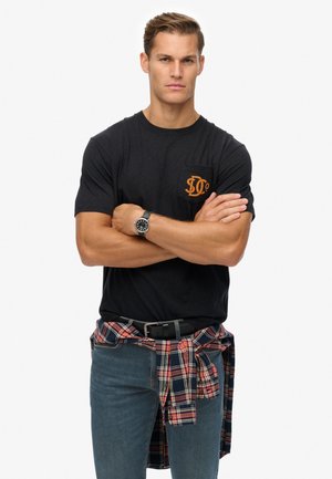 Zwarte katoenen T-shirt met oranje zaklogo, gecombineerd met een blauwe spijkerbroek en een rode geruite shirt knoop om de taille. RVS horloge.