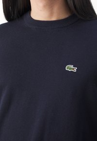 Mörkblå stickad tröja med rund halsringning, med en liten grön krokodil-logotyp på bröstet. Tyget ser mjukt och slätt ut.