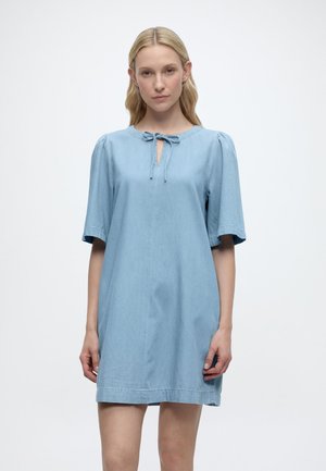 Blonde Frau trägt ein locker sitzendes, hellblaues kurzärmeliges Kleid mit einer vorderen Schnürung am Ausschnitt, steht vor einem weißen Hintergrund.