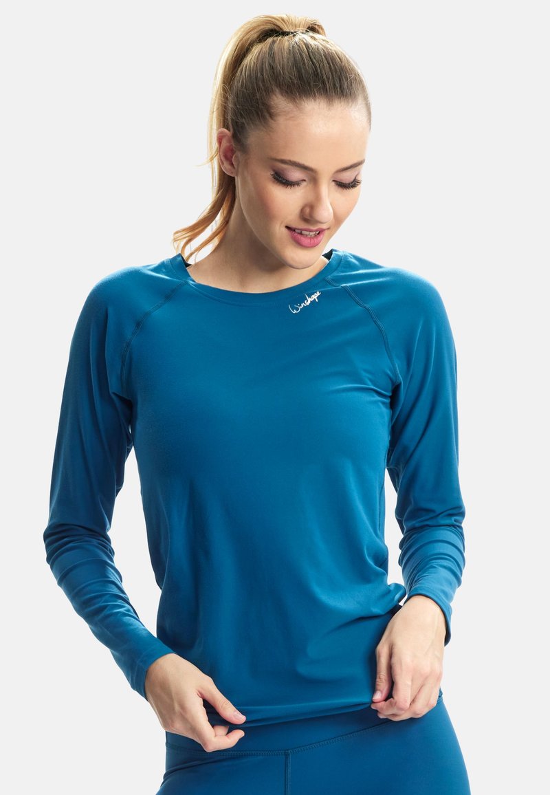 Kobieta z blond kucykiem patrząca w dół, ubrana w dopasowaną, długorękawową sportową bluzkę w kolorze morskim oraz dopasowane legginsy w tym samym kolorze na białym tle.