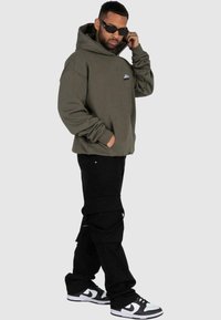 Felpa verde oliva con tasca frontale e logo, abbinata a pantaloni cargo neri con tasche laterali e sneakers bianco e nero.